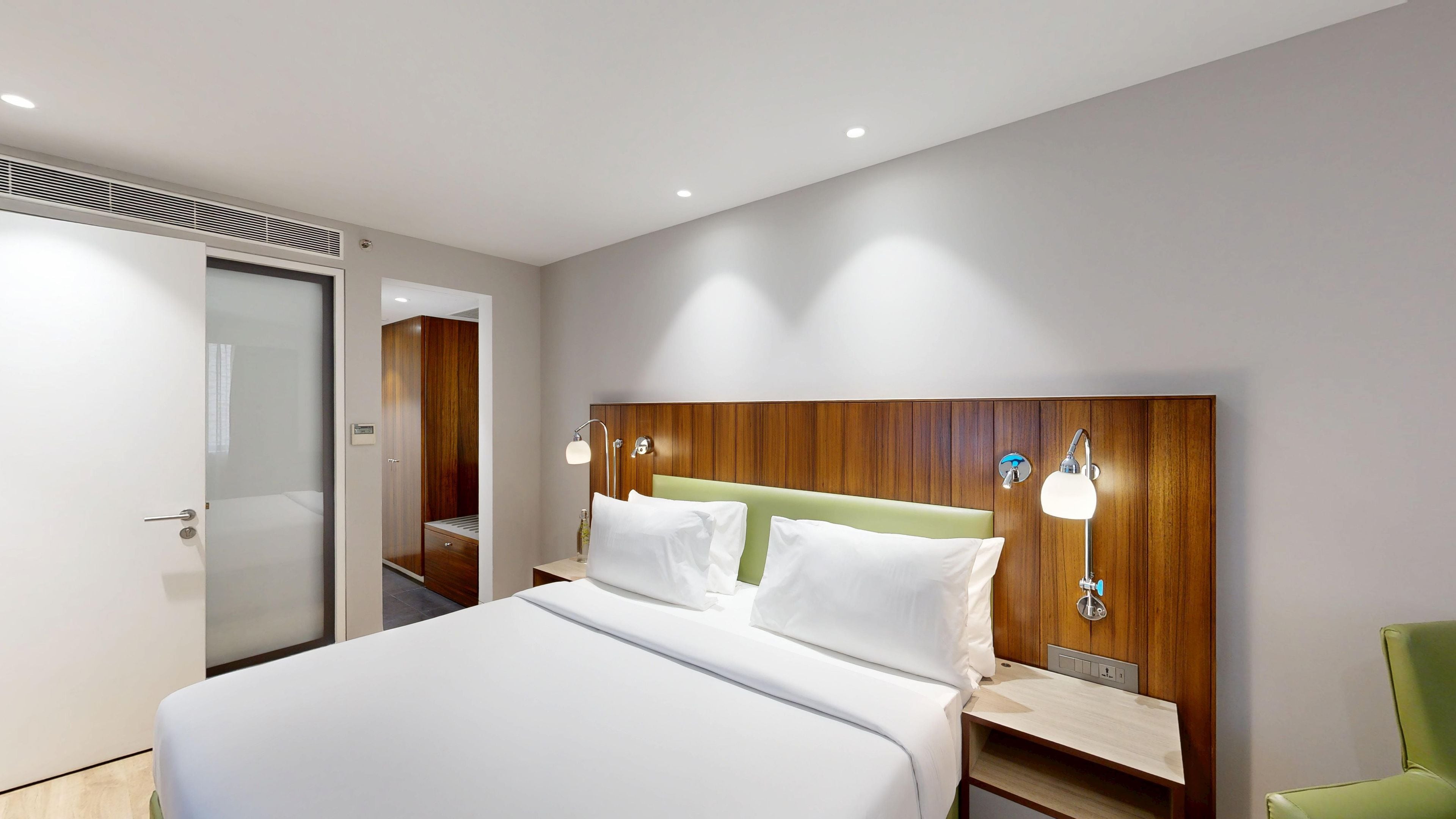 Une chambre d’hôtel moderne avec un lit, des tables de chevet, un fauteuil vert et une tête de lit en bois.