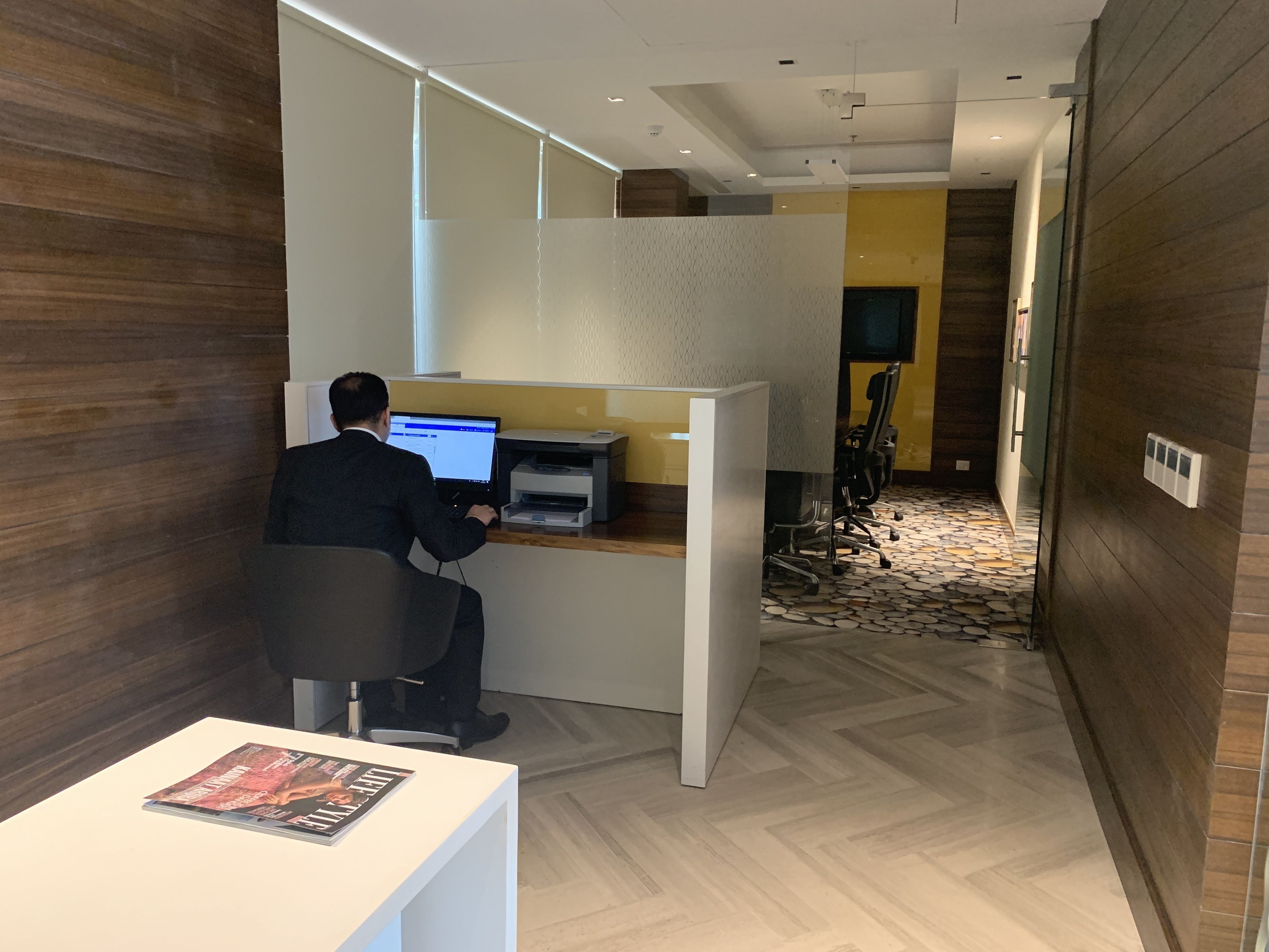 A man working at a desk with a computer and printer, in a modern office with wooden walls and a patterned floor.