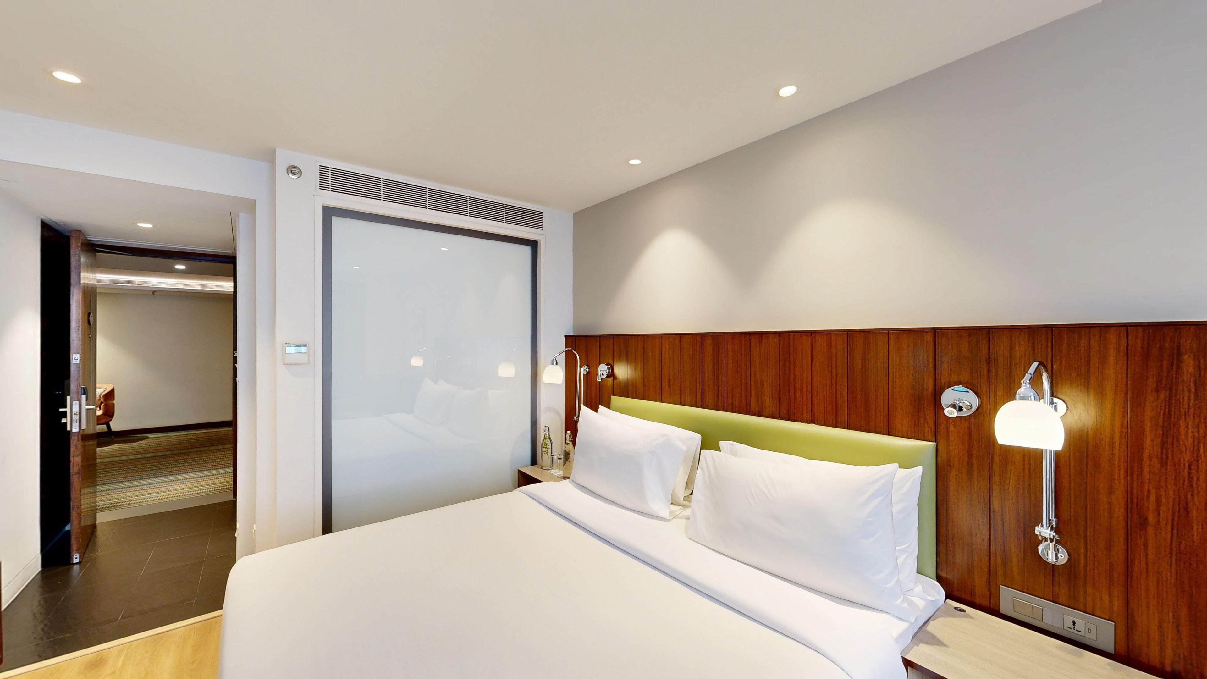 A hotel room with a large bed, bedside lamps, a wooden headboard, and a door leading to another room.