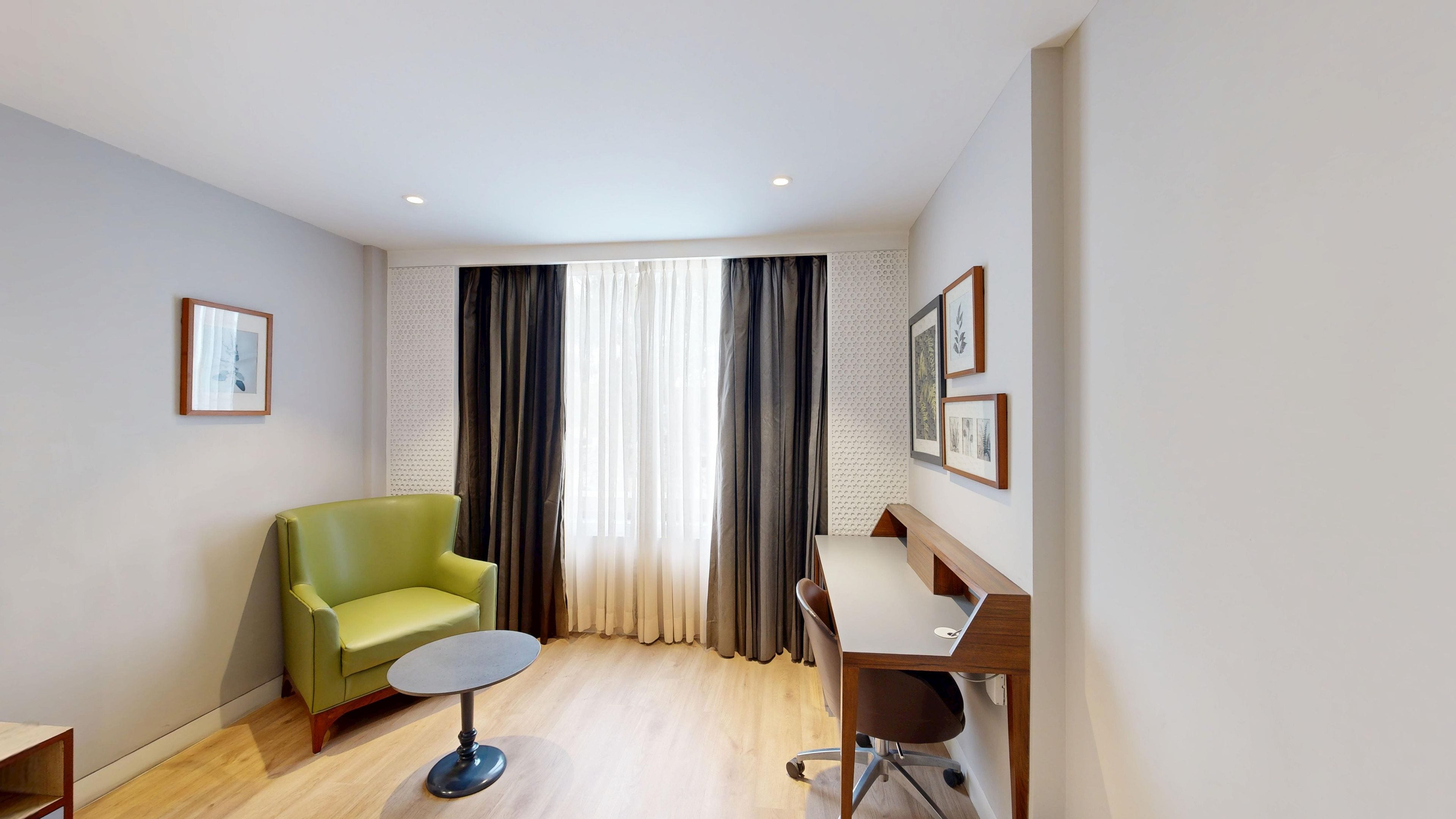 A room with a green armchair, a small round table, a wooden desk, and a large window with curtains.