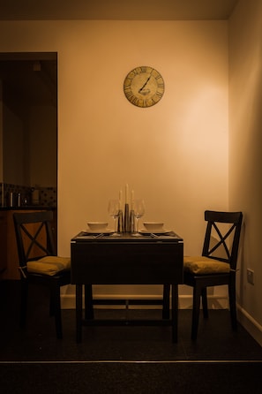 A dining area with a table set for two, a wall clock, and a kitchen in the background.