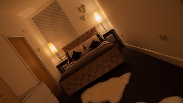 A bedroom with a tufted headboard, two bedside lamps, a bed with a white furry rug, and a window with blinds. - Station Suite - Simple2let Serviced Apartments (Halifax)