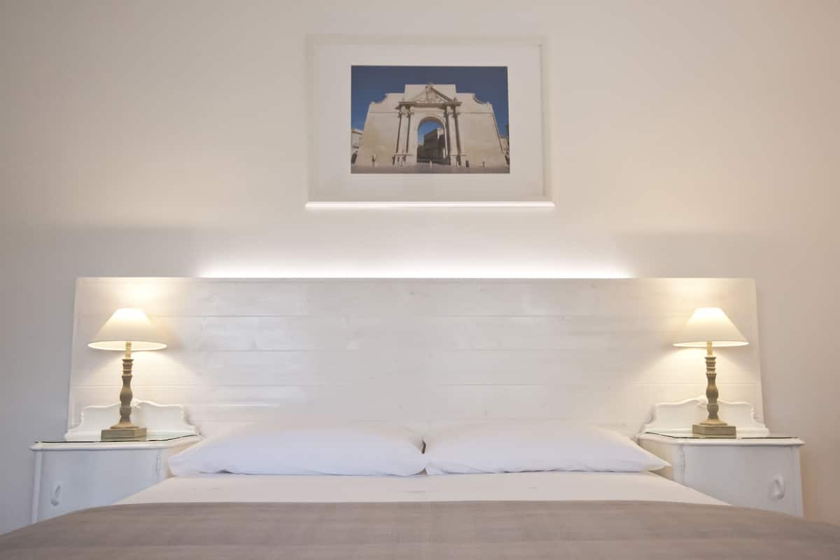 Une chambre à coucher avec une tête de lit blanche, deux lampes de chevet, un tableau encadré au-dessus de la tête de lit et un lit avec une literie blanche.