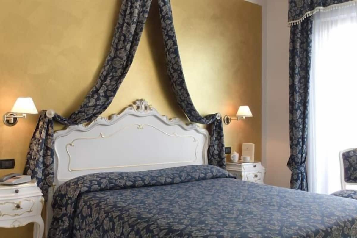 Een bed met een witte hoofdbord en beddengoed met bloemenprint, aan weerszijden geflankeerd door twee nachtkastjes met lampen, in een kamer met een gele wand en gordijnen met een patroon.