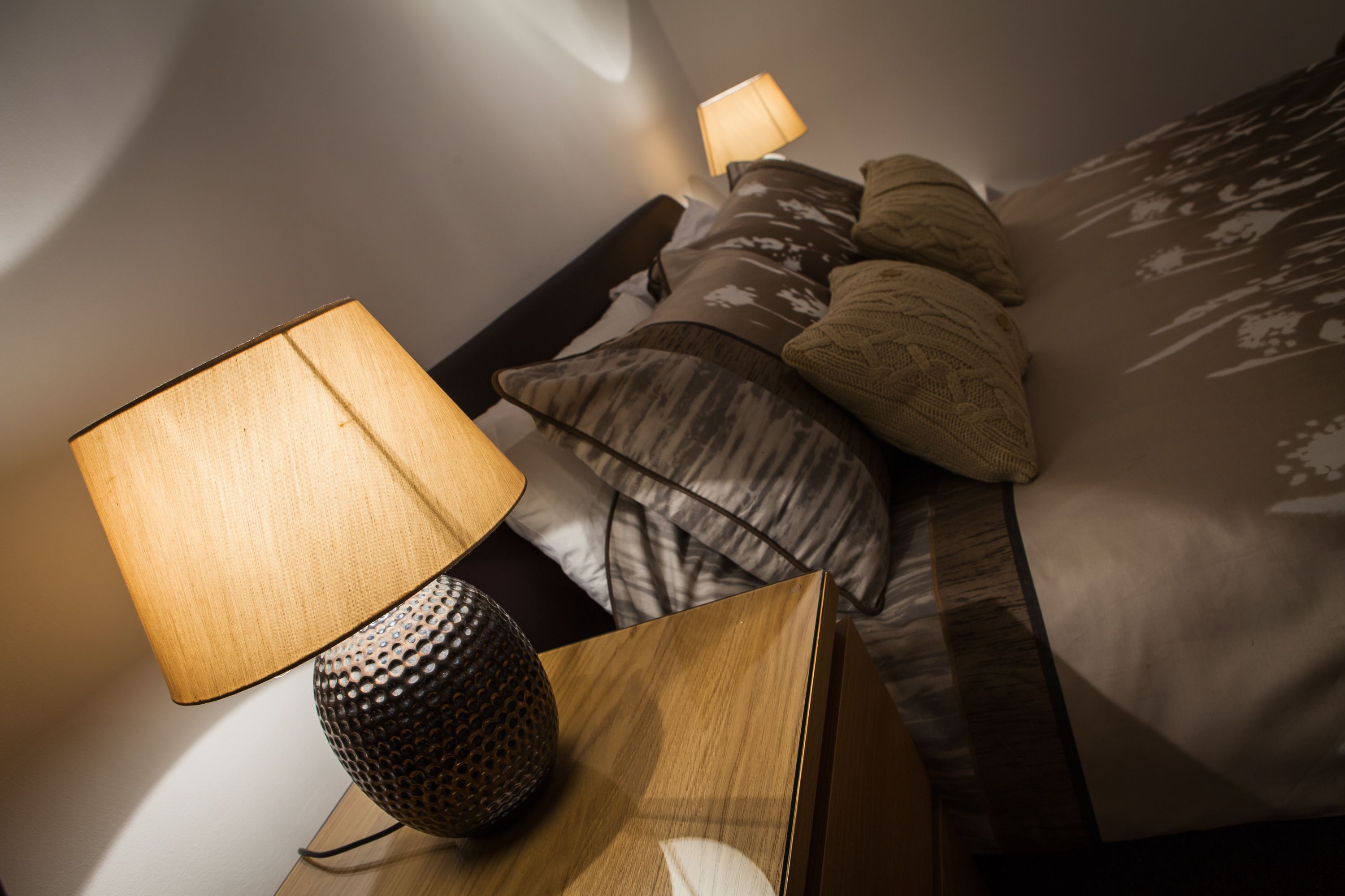 A neatly made bed with patterned bedding, a wooden bedside table with a lamp, and a wall-mounted reading light.
