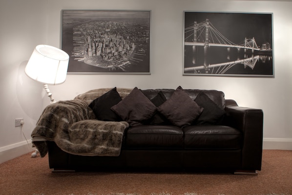 A living room with a black leather sofa, a throw blanket, and two framed cityscape photographs on the wall.