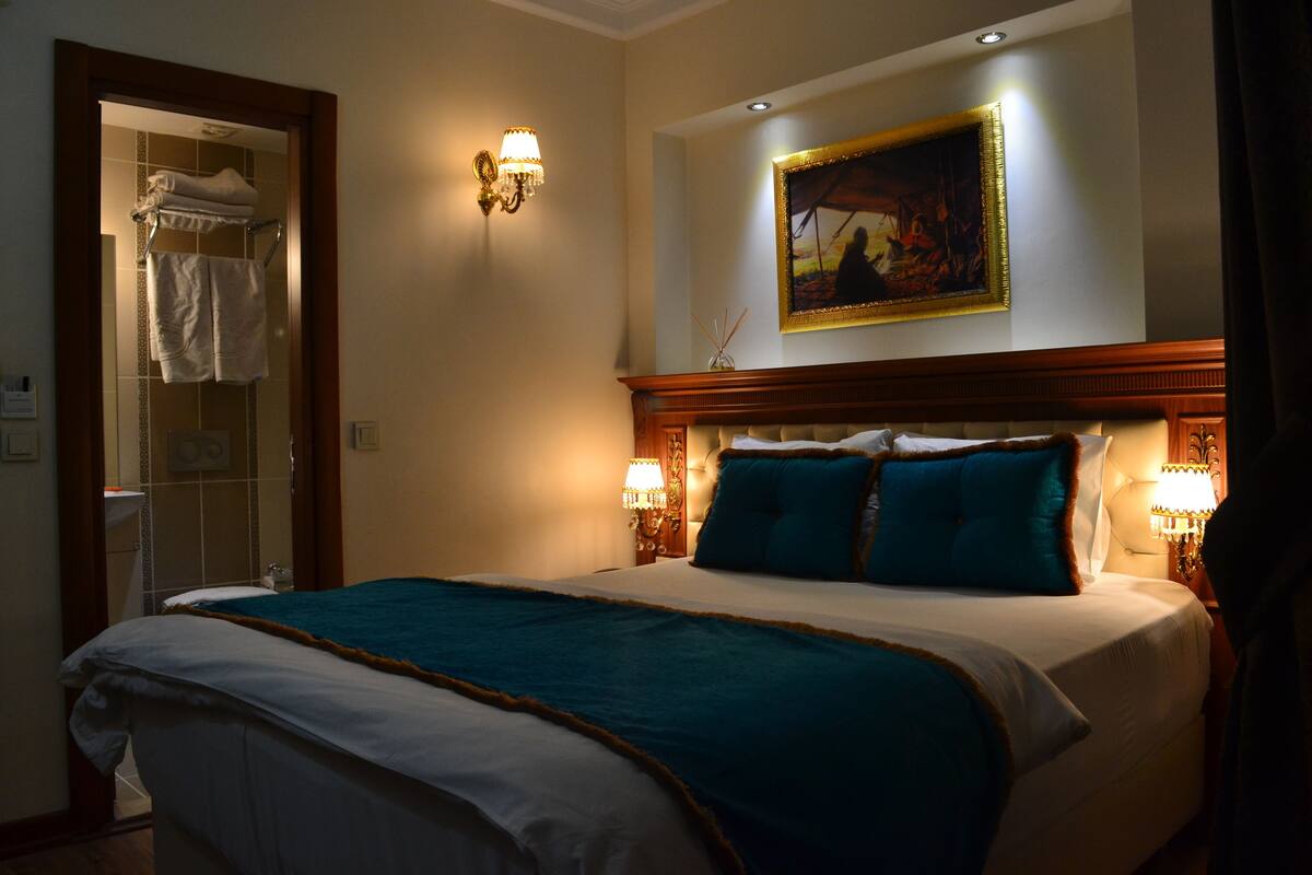A hotel room with a large bed, two wall-mounted lamps, a painting above the bed, and a bathroom with a towel rack.