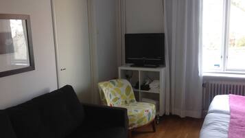 Una habitación con un sofá negro, una silla estampada con hojas, un televisor sobre un mueble bajo y una cama con una manta rosa.