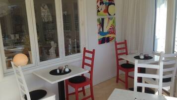 Una habitación con paredes blancas, un ventanal grande y un cuadro abstracto colorido. Hay dos mesas blancas con sillas rojas, y una mesita redonda con camino de mesa negro.