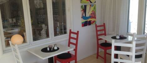 Una habitación con paredes blancas, un ventanal grande y un cuadro abstracto colorido. Hay dos mesas blancas con sillas rojas, y una mesita redonda con camino de mesa negro.
