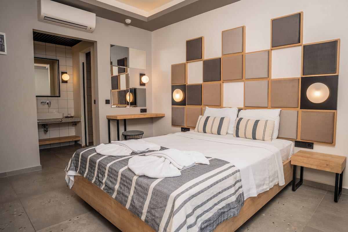 A modern hotel room with a large bed, a bedside table, a vanity with a mirror, and a wall-mounted light fixture.