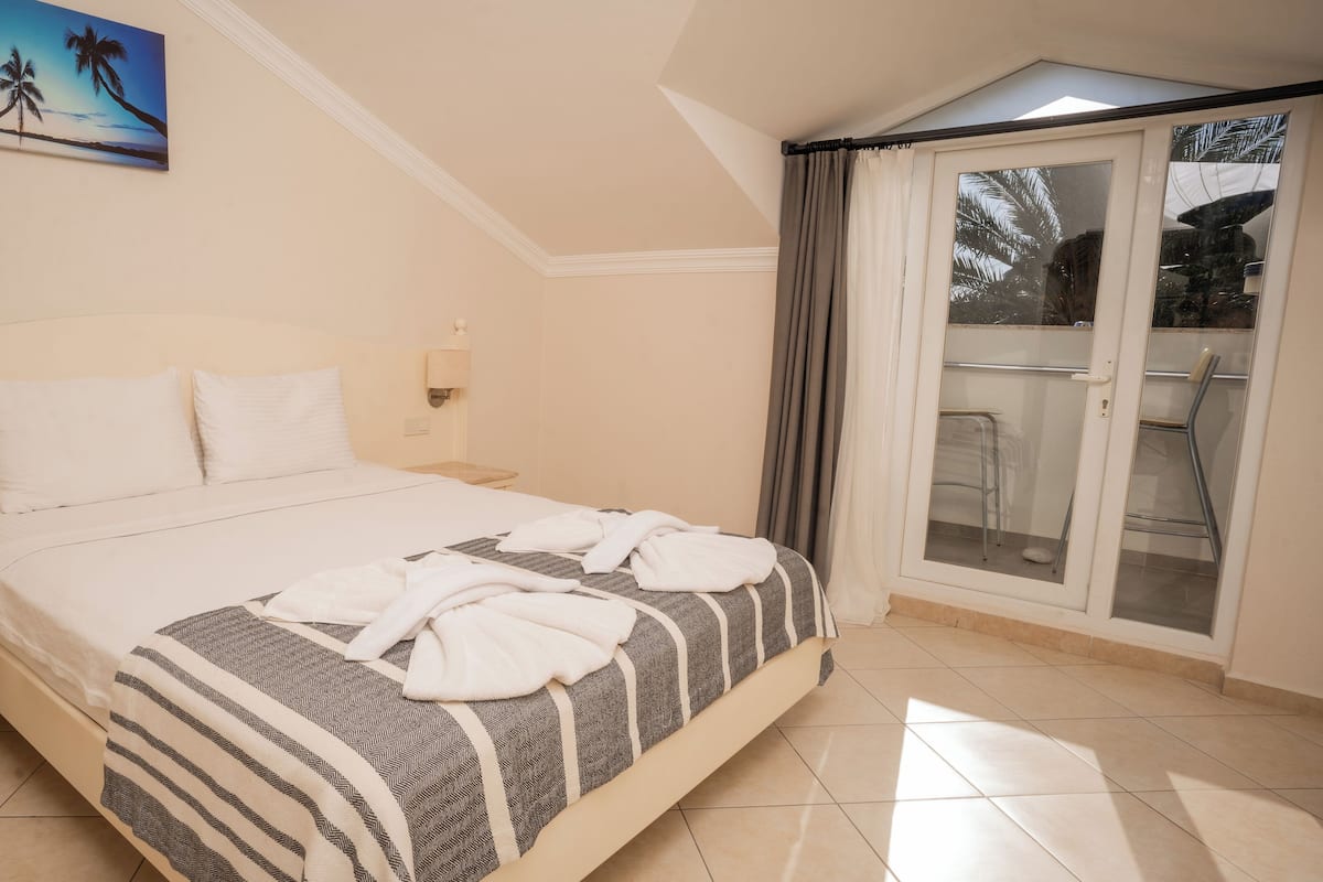 A bedroom with a bed, a nightstand, a wall-mounted lamp, a window with curtains, and a view of the outdoors.
