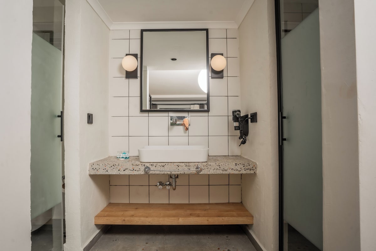 Un baño con lavamanos blanco, un espejo, dos luces empotradas en la pared y un perchero para toallas.