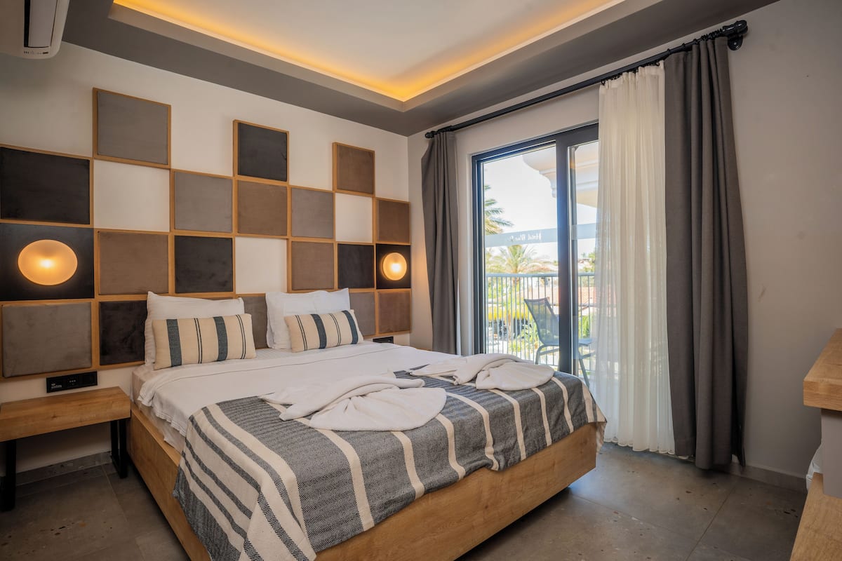 A modern hotel room with a large bed, a bedside table, a wall-mounted shelf, and a window with curtains.