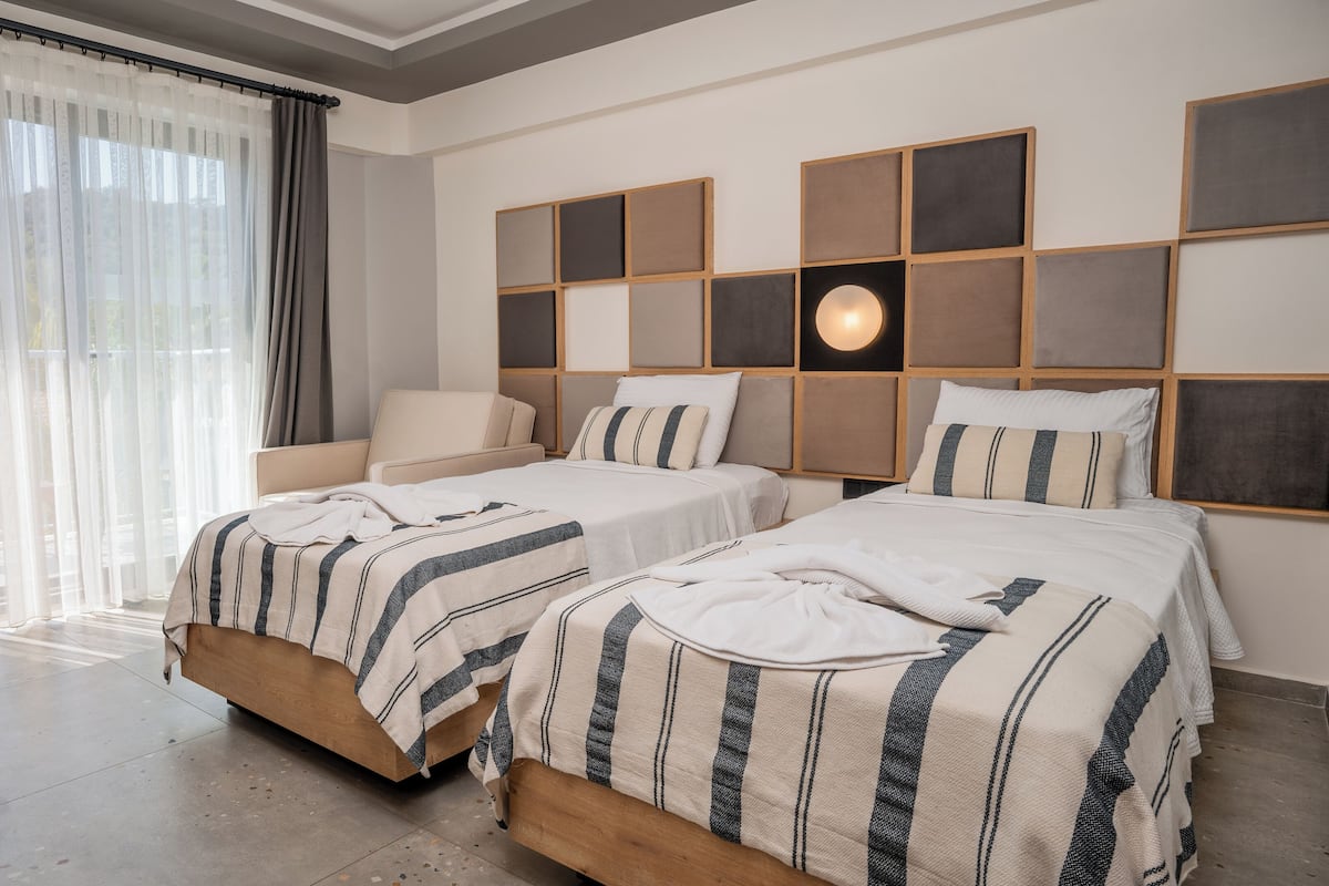A hotel room with two beds, a sofa, a large window with sheer curtains, and a wall with a geometric light fixture.