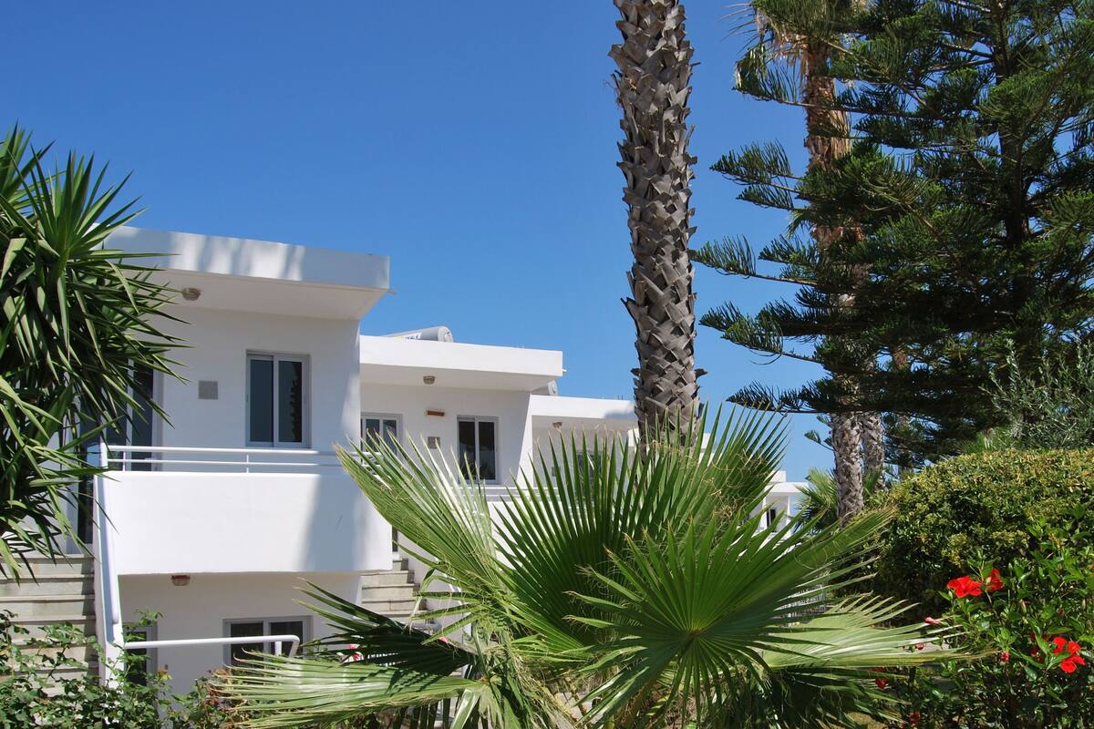Um edifício moderno branco com varanda, rodeado por palmeiras e flores vermelhas.
