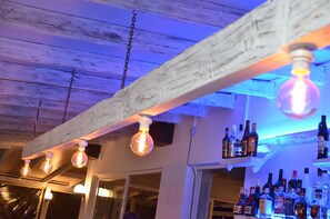 A bar area with exposed wooden beams, hanging light bulbs, and shelves stocked with liquor bottles.