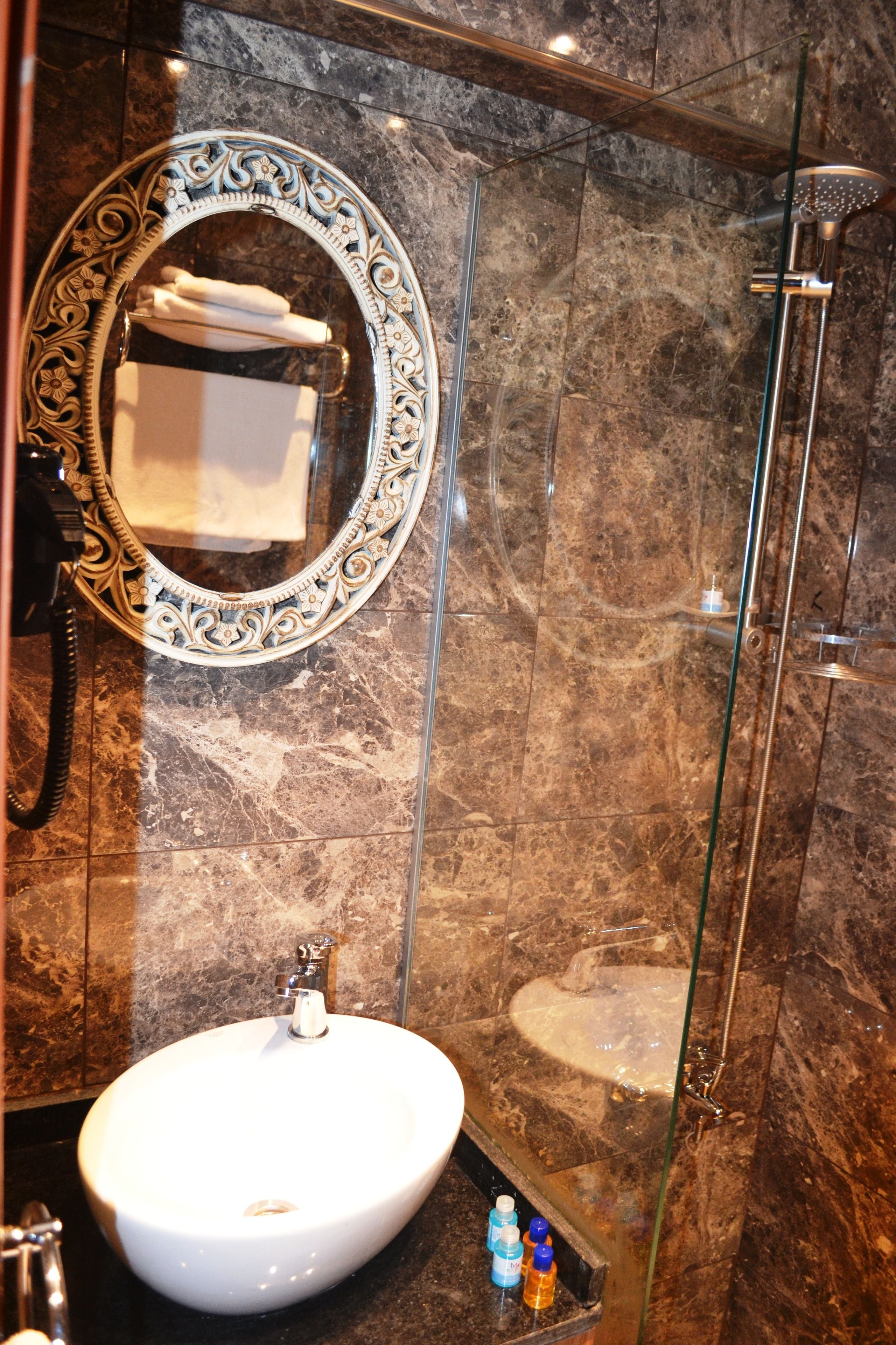 A bathroom with a marble wall, a round mirror with a decorative frame, a white sink, and a glass-enclosed shower.