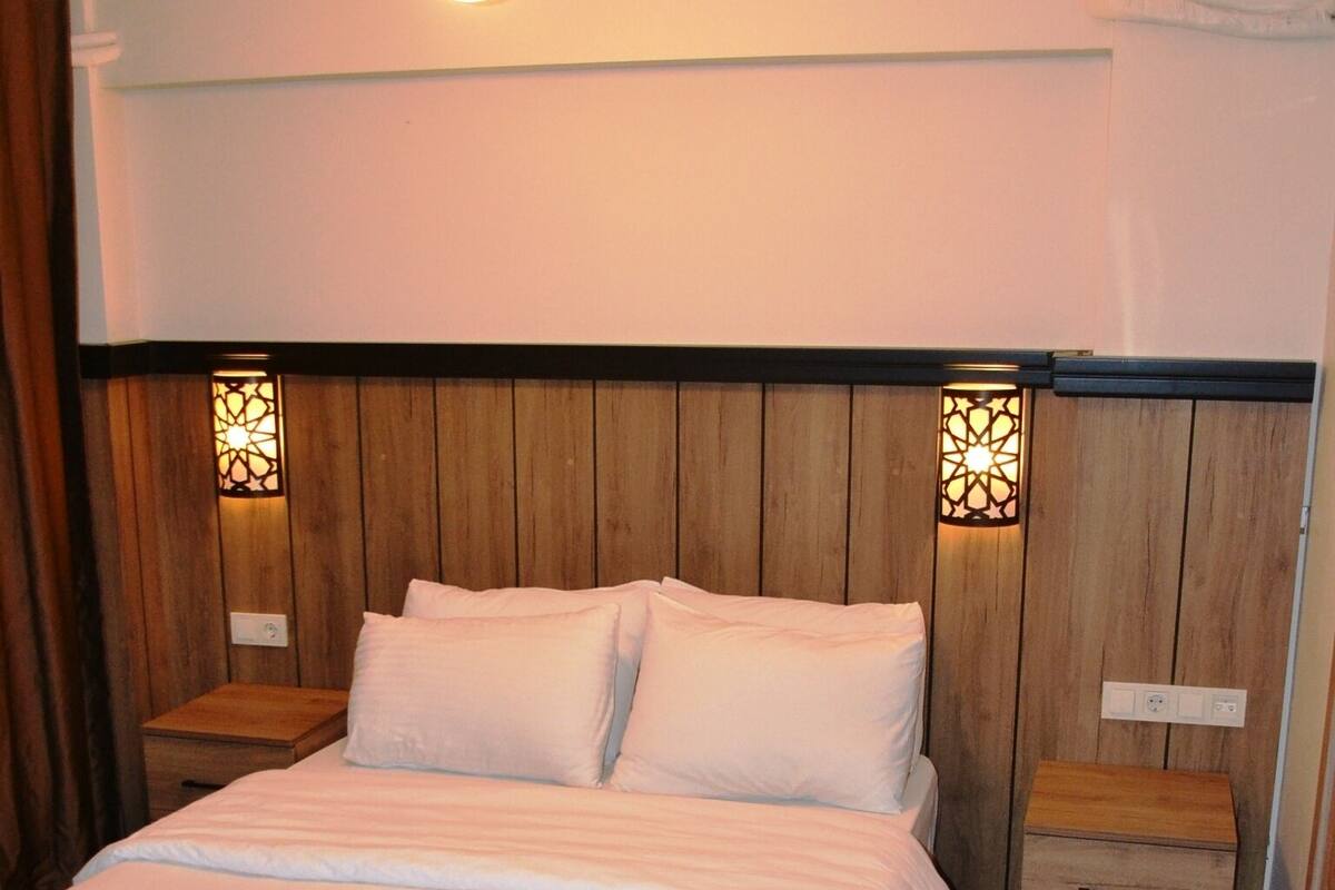 A bedroom with a wooden headboard, a large bed with white bedding, two bedside tables, and wall-mounted sconces.