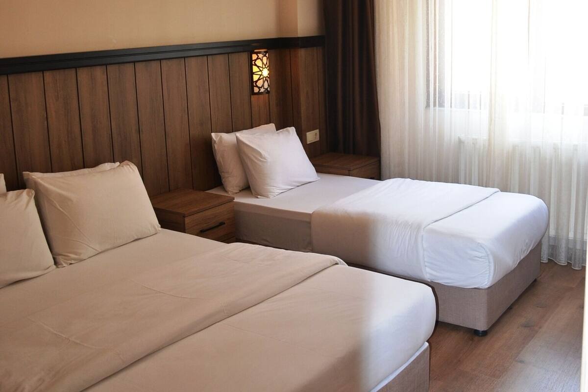 A hotel room with two beds, wooden headboard, a small nightstand, a wall-mounted light fixture, and a window with sheer curtains.