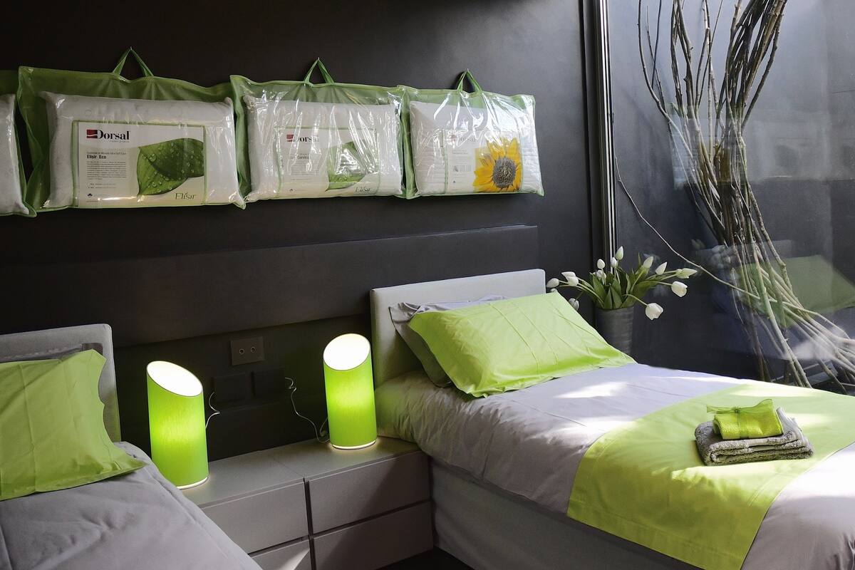 Une chambre d’hôtel avec un lit, des oreillers verts et une fenêtre donnant sur des branches d’arbres. On peut également voir des sacs verts accrochés au mur.