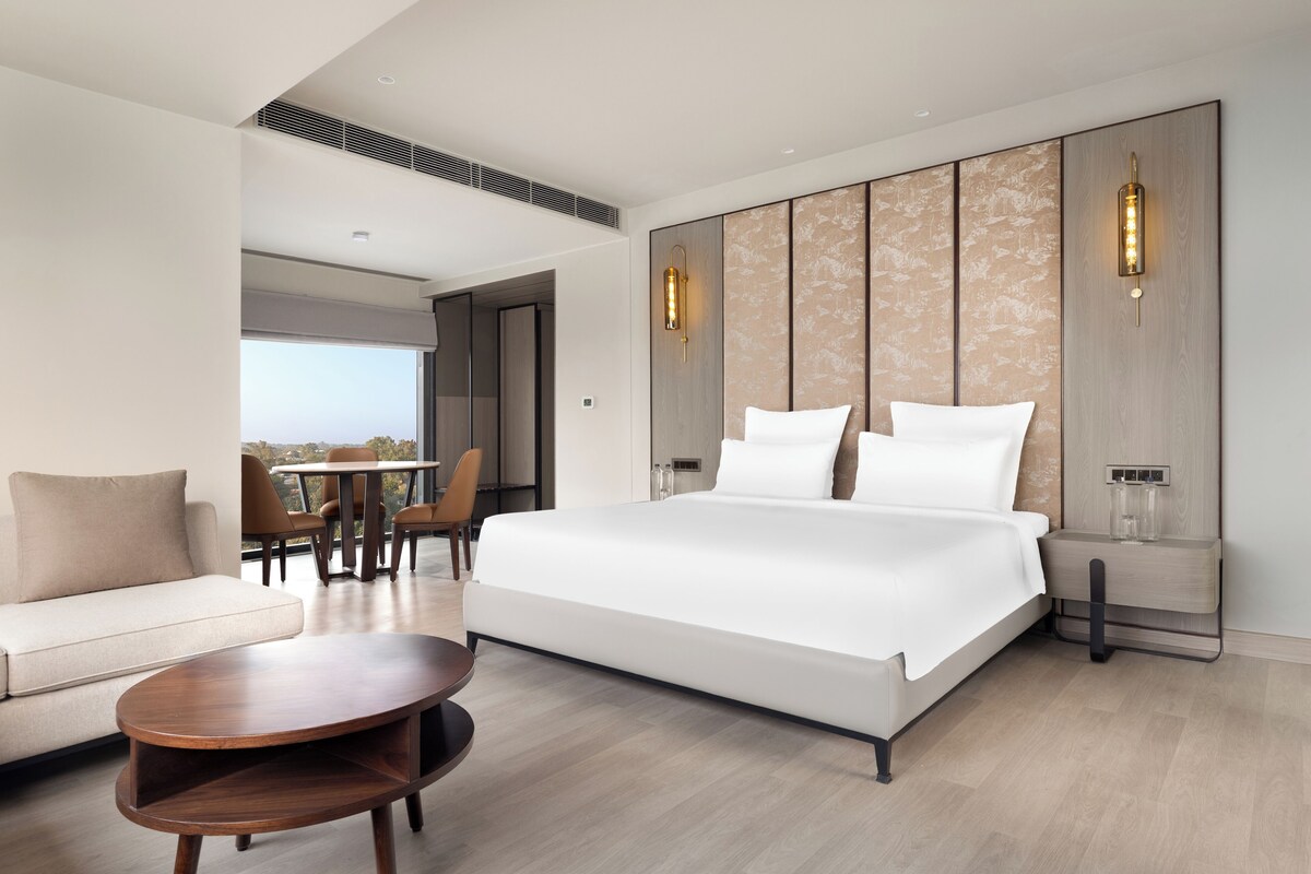 A modern hotel room with a large bed, a sofa, a dining table, and two wall-mounted lamps.