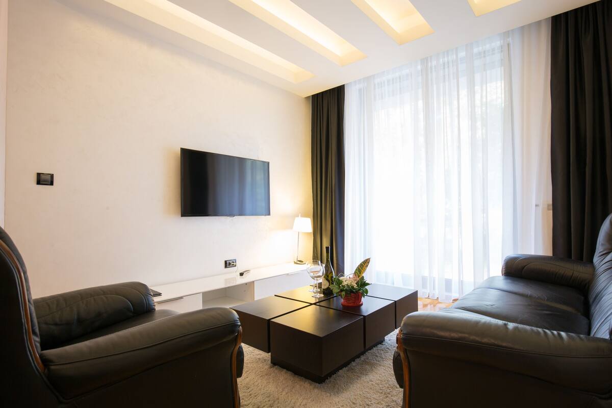 Uma sala de estar moderna com televisão de tela plana na parede, um sofá escuro, uma mesa de centro com uma planta e uma janela grande com cortinas leves.