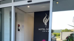 A modern hotel entrance with a glass door, a sign reading "HOTEL IVON", and a view of the surrounding landscape.