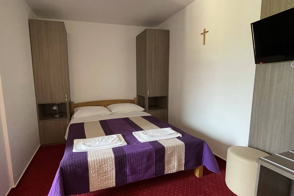A small, neatly arranged bedroom with a bed, bedside tables, a television, and a cross on the wall.