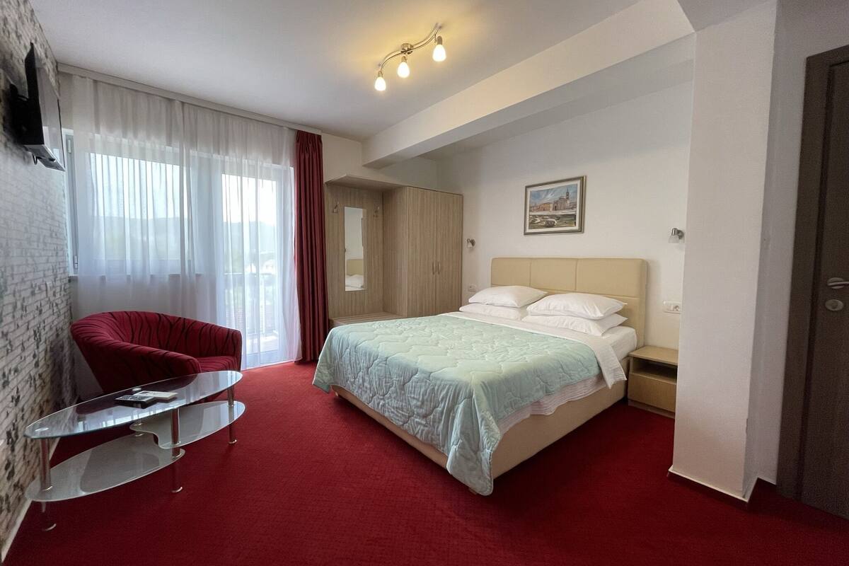 A hotel room with a bed, a red chair, a small table, and a window with curtains.
