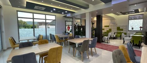 Un comedor moderno con varias mesas y sillas, ventanas amplias que ofrecen una vista, y un techo bien iluminado con luces empotradas.