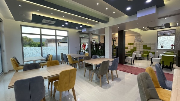 A modern dining area with multiple tables and chairs, large windows with a view, and a well-lit ceiling with recessed lighting.