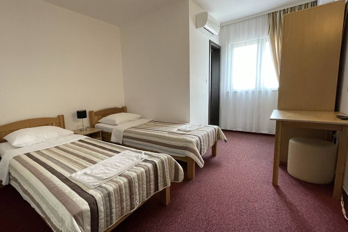 A hotel room with two single beds, a desk, a chair, a lamp, and a window with curtains.