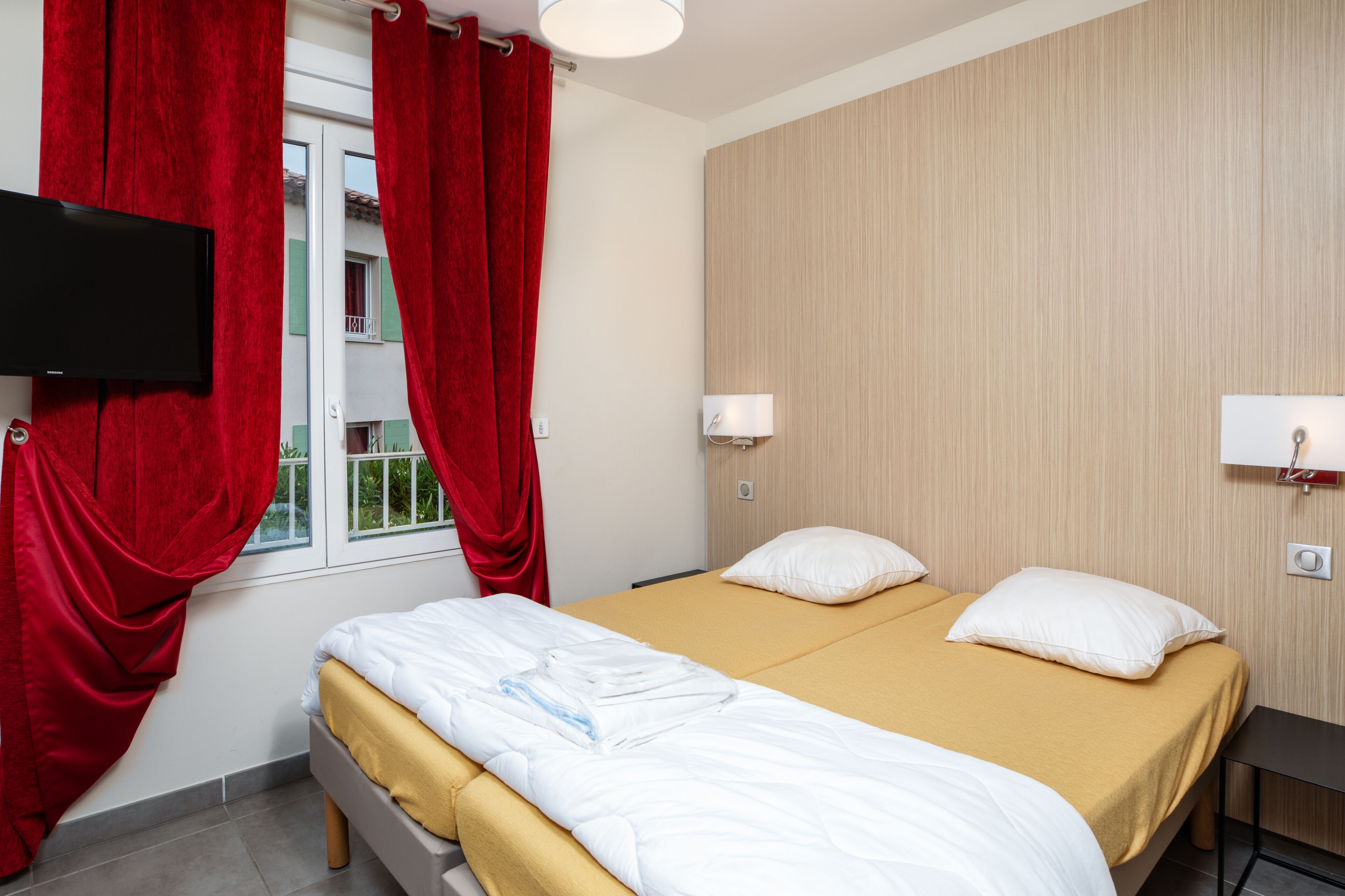 A hotel room with two beds, red curtains, a TV, a bedside table, and a window with a view of a building.