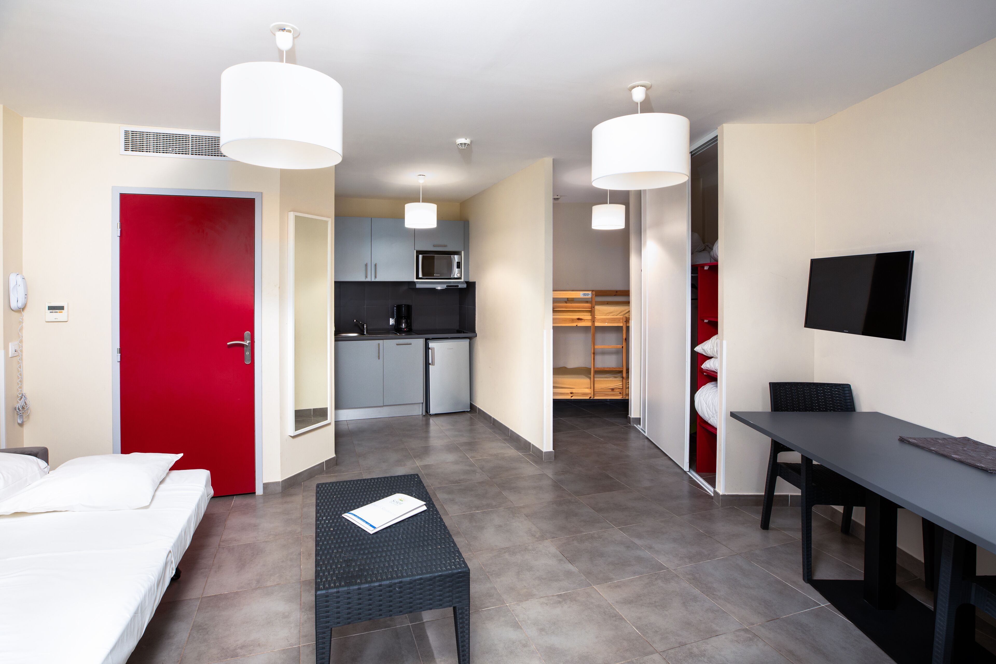 Et moderne hotelværelse med seng, lille bord, skrivebord, fjernsyn, tekøkken og en rød dør.