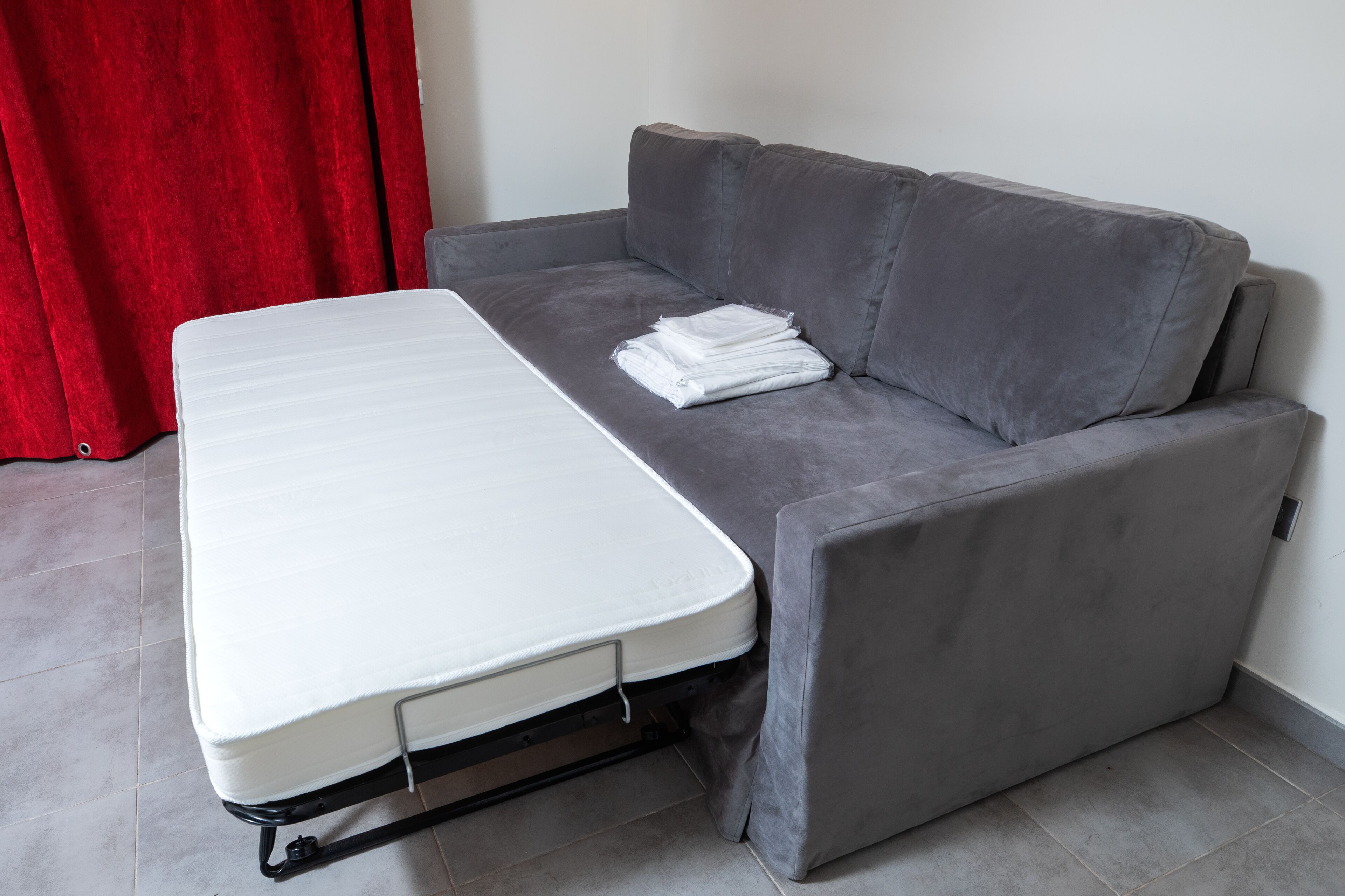 A compact room with a grey sofa and a white mattress on wheels, suggesting a convertible sofa-bed setup.