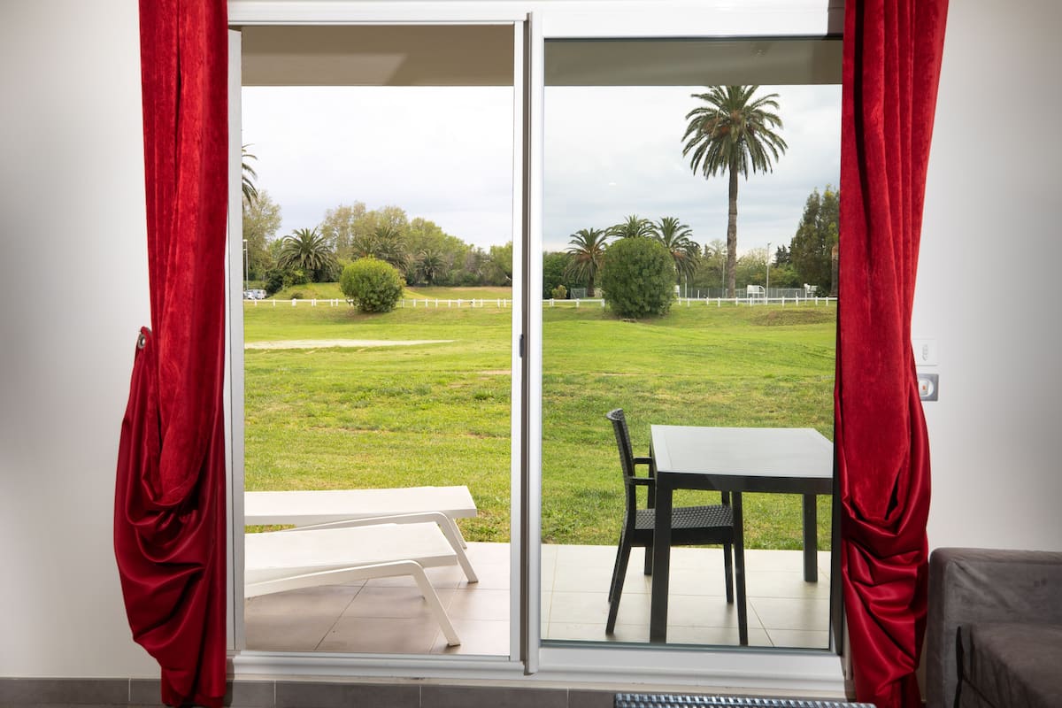 Une pièce dotée d’une porte-fenêtre coulissante donnant sur une terrasse avec une table et des chaises, un banc blanc et une vue sur un espace verdoyant agrémenté d’arbres et d’un palmier.