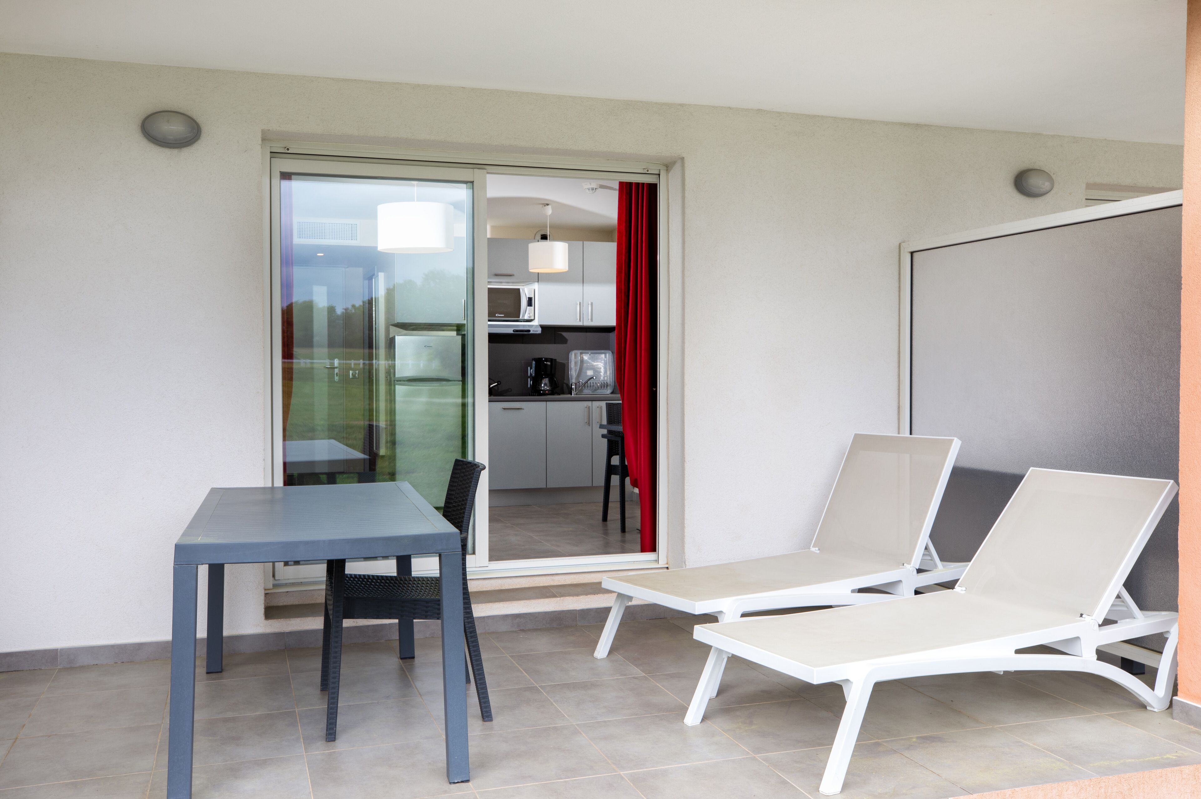 Un balcón con mesa y sillas, una cocina visible a través de una puerta abierta y una cortina roja.