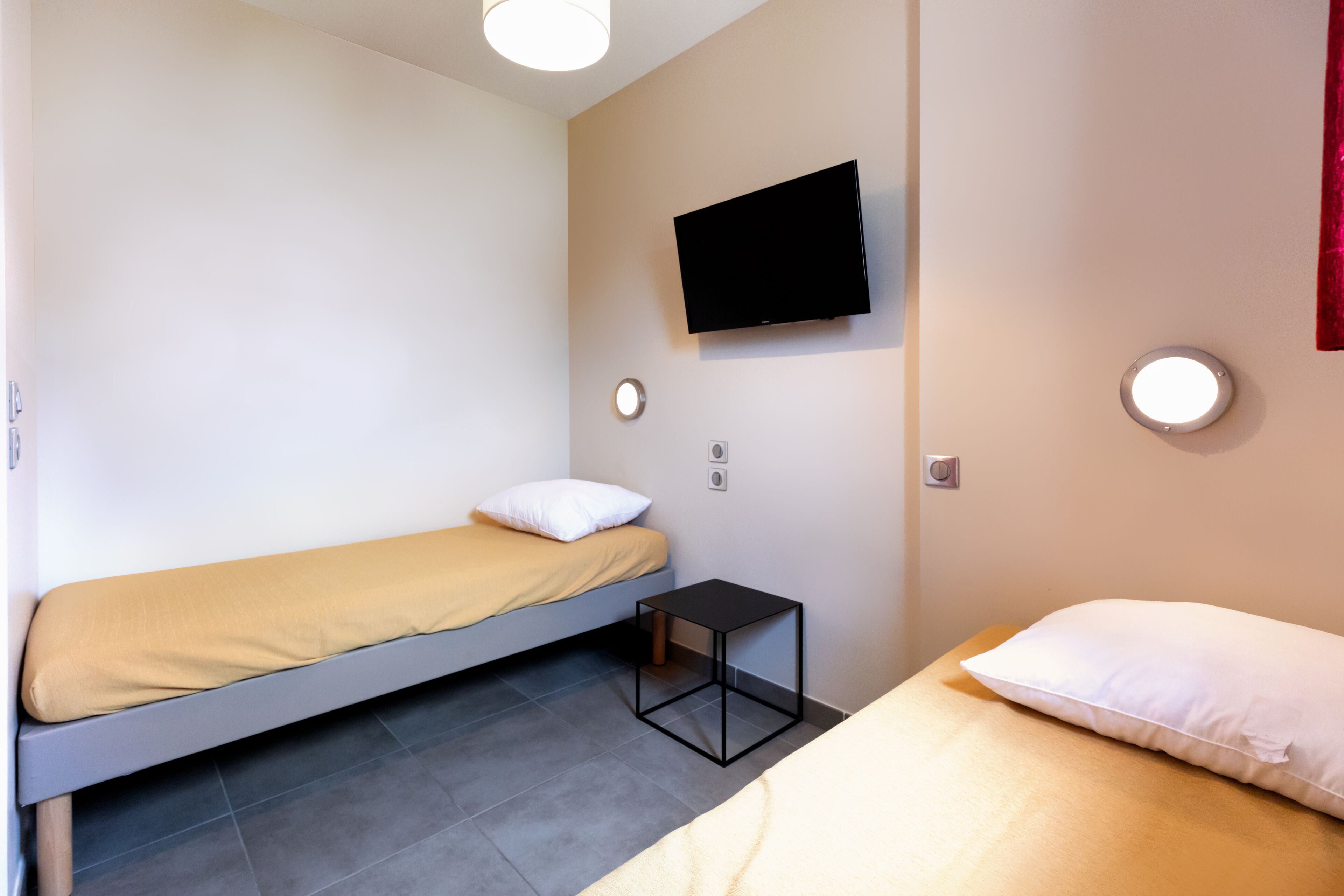 A small, modern room with two single beds, a flat-screen TV mounted on the wall, and a small table between the beds.