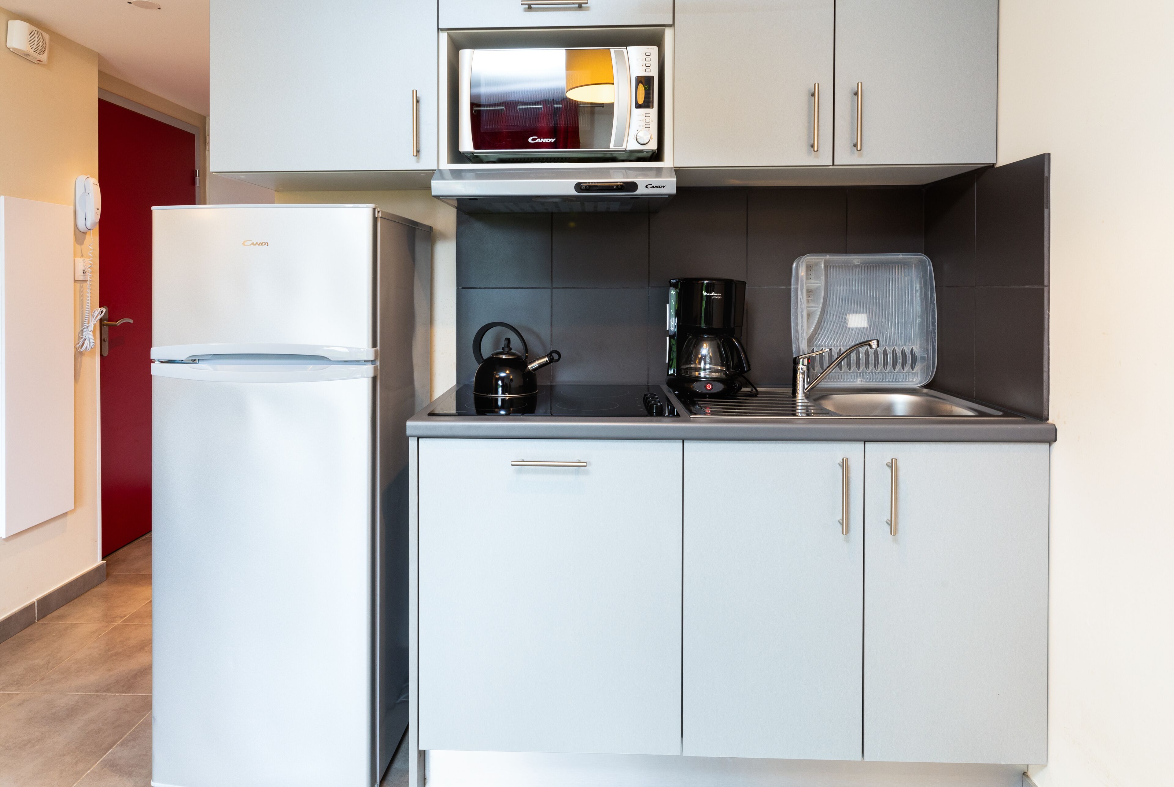 Een compacte keuken met een witte koelkast, een magnetron, een waterkoker en een koffiezetapparaat.