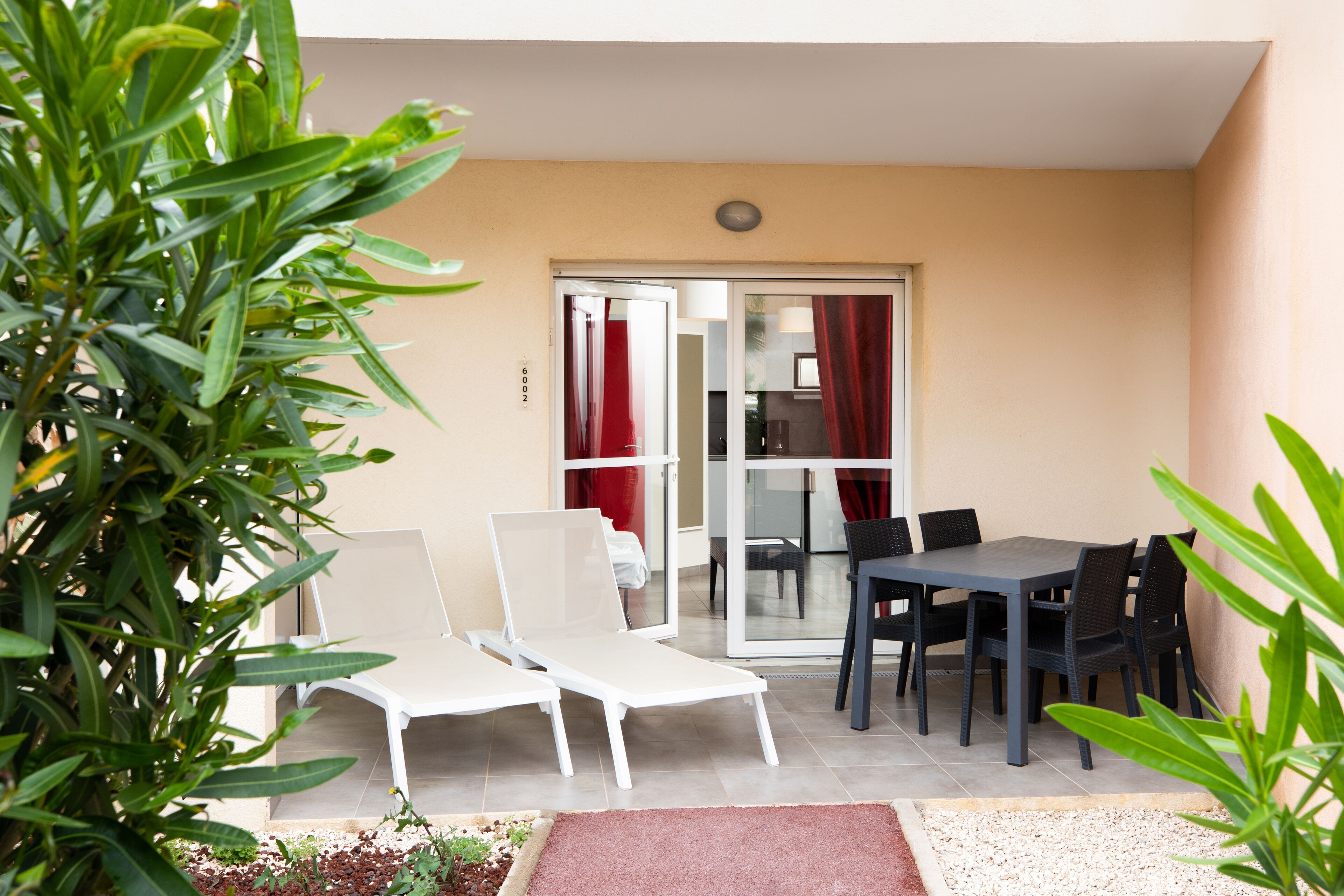 Un patio con dos sillones blancos, una mesa de comedor con sillas y un ventanal corredero que da a una habitación.