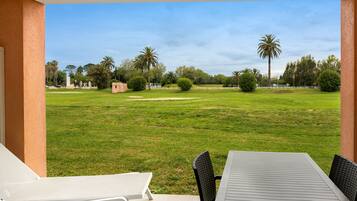An outdoor patio with a table and chairs, a view of a well-maintained lawn with palm trees, and a clear sky.