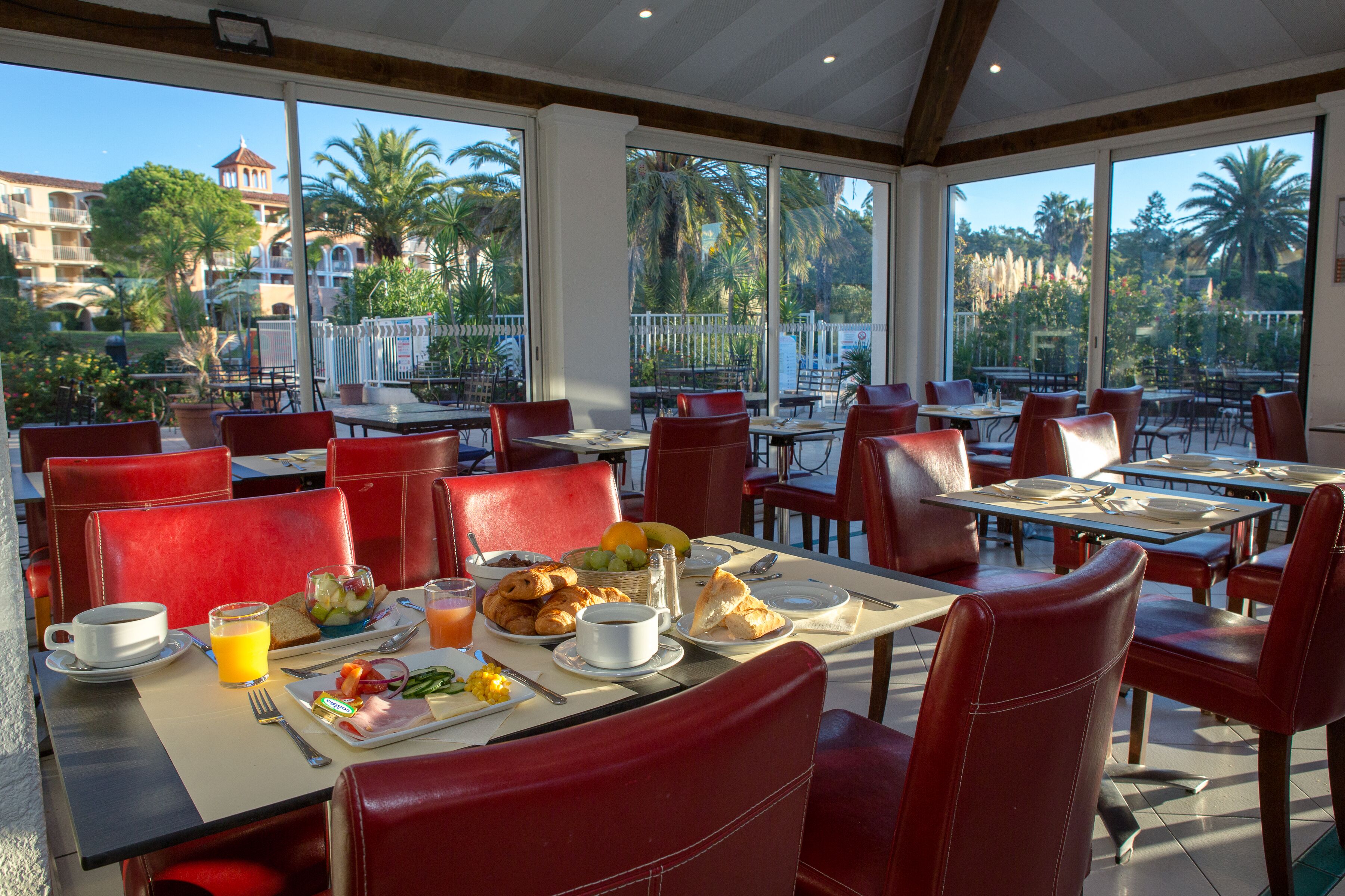 Comedor con sillas y mesas rojas, alimentos para el desayuno y vistas a palmeras y edificios.