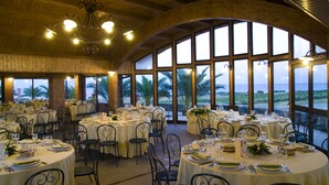 A restaurant with round tables set for dining, large windows with a view of palm trees, and a wooden ceiling. - Serra San Biagio (Calatabiano)