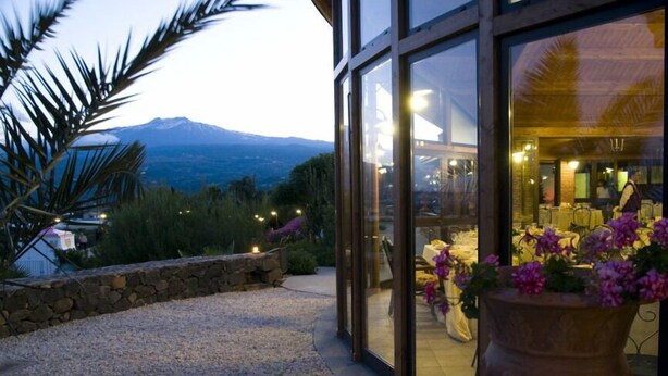 A modern building with large glass windows, a stone retaining wall, and a view of a mountain range. - Serra San Biagio (Calatabiano)