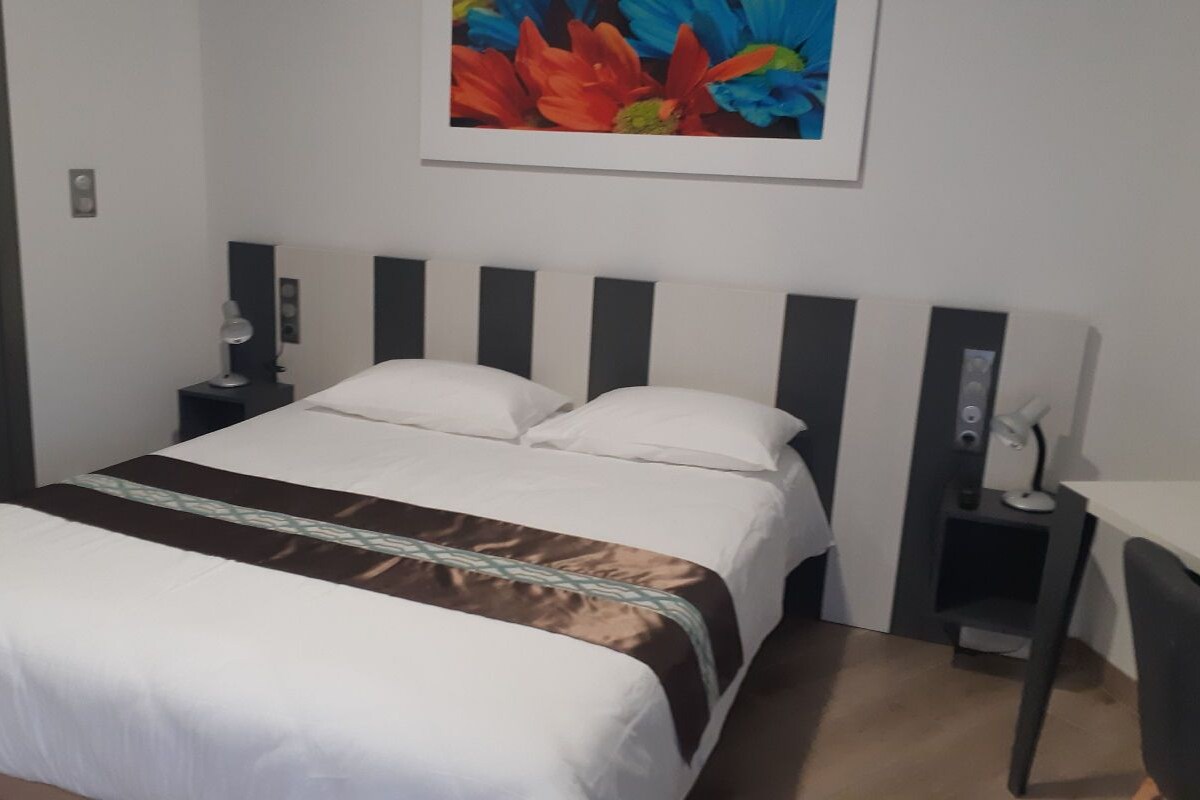 A hotel room with a bed, a desk, a chair, a nightstand, and a wall-mounted lamp.