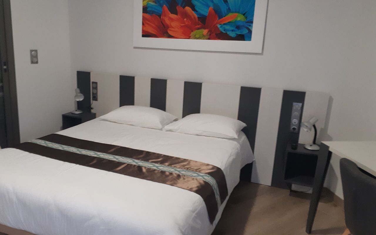 A hotel room with a bed, a desk, a chair, a nightstand, and a wall-mounted lamp.