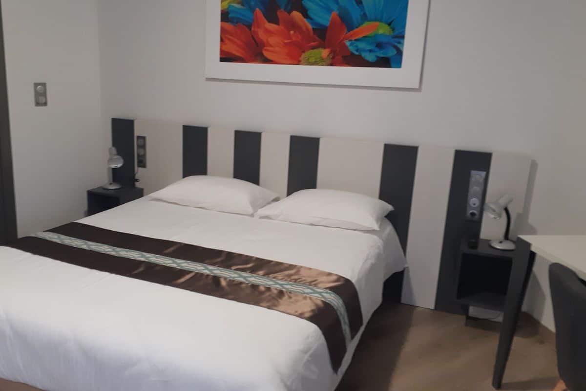 A hotel room with a bed, a desk, a chair, a nightstand, and a wall-mounted lamp.