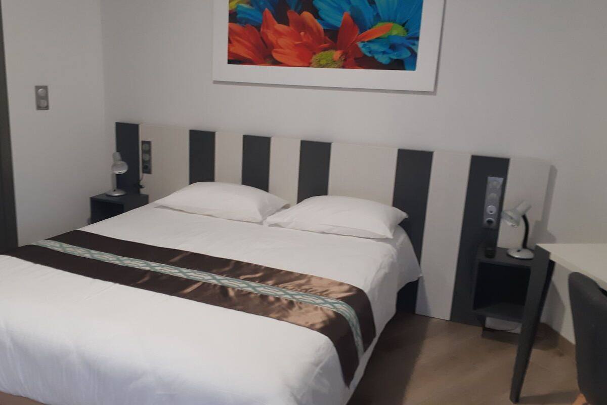 A hotel room with a bed, a desk, a chair, a nightstand, and a wall-mounted lamp.