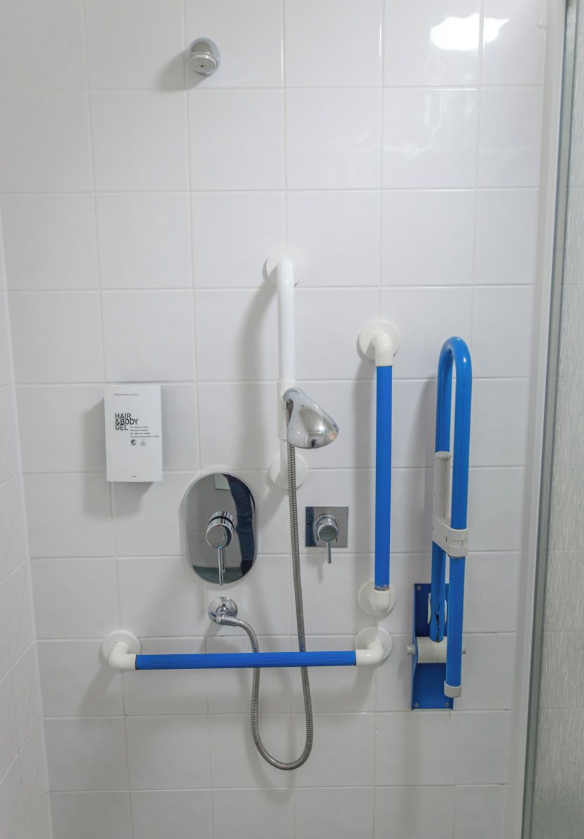 A bathroom with a white tiled wall, a handheld showerhead, a handheld shower attachment, a handheld shower hose, a blue grab bar, and a white soap dispenser.